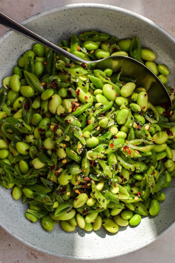 Close up of a edamame bean and sugar snap pea salad with dried red chilli flakes and black sesame seeds sprinkled on top.