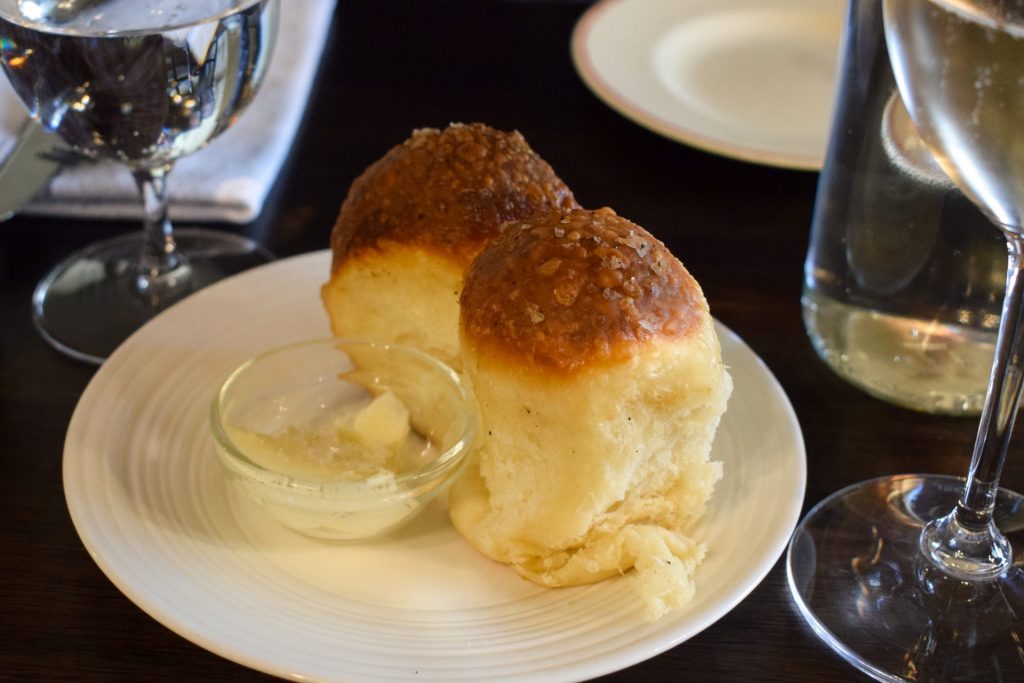 A pair of milk buns with golden tops and a glass dish of butter on a white plate.