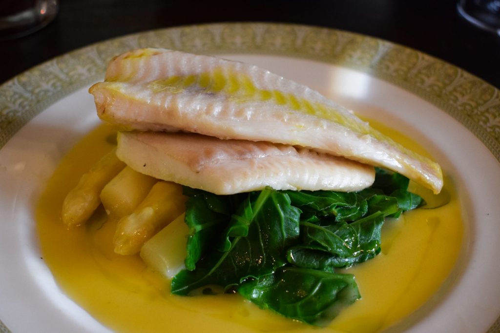 Two fillets of poached plaice on top of a pile of sea beet leaves and white asparagus sitting in a puddle of beurre blanc.