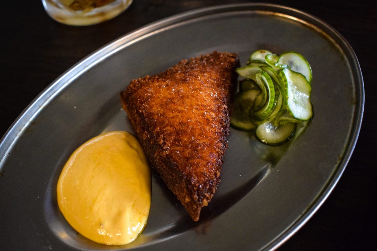 A triangle of scallop toast with a dollop of seafood emulsion and pile of pickled cucumber slices on a metal plate.