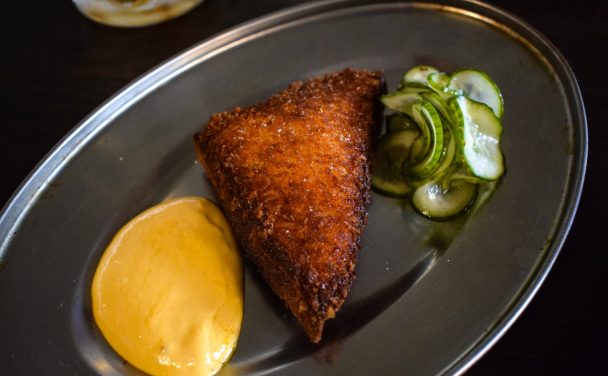 A triangle of scallop toast with a dollop of seafood emulsion and pile of pickled cucumber slices on a metal plate.