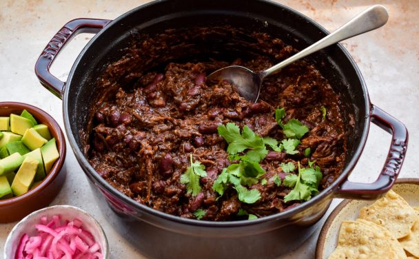 Side on view of beef chili in a burgundy casserole dish. Pots of avocado, pickled red onions and tortilla chips are around the pot.