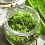Side on view of an open jar of wild garlic salt with wild garlic leaves off to one side.