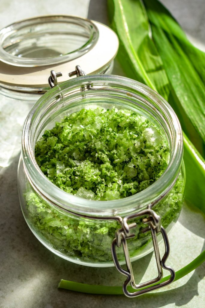 Side on view of an open jar of wild garlic salt with wild garlic leaves off to one side.