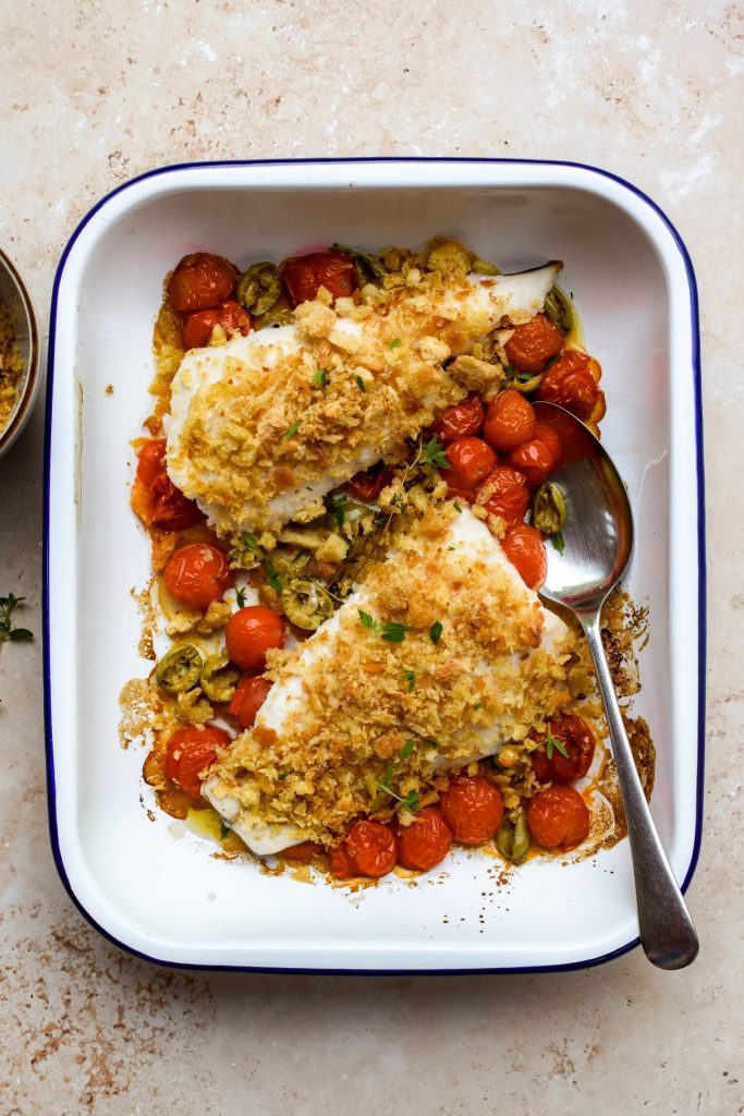 Overhead image of two hake fillets topped with breadcrumbs in a tray with cherry tomatoes and olives.