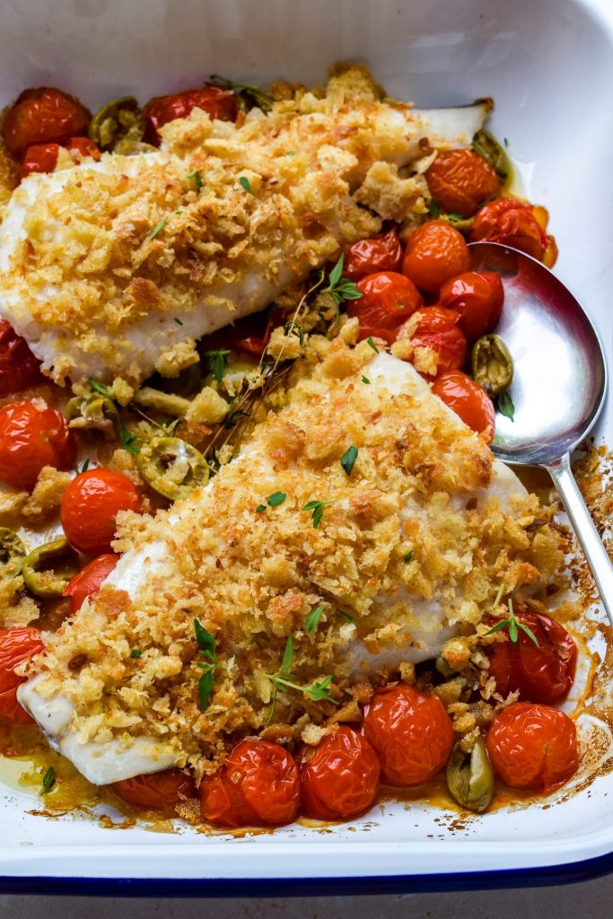 Close up of a tray of breadcrumb-baked hake with cherry tomatoes and torn green olives.