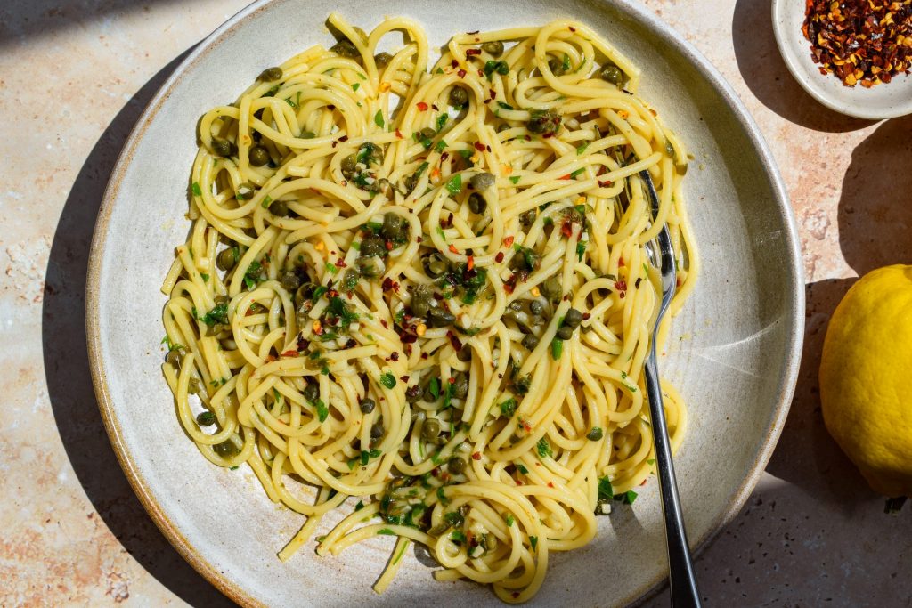 Overhead image of a bowl of caper, lemon, garlic and chilli spaghetti in direct sunlight.
