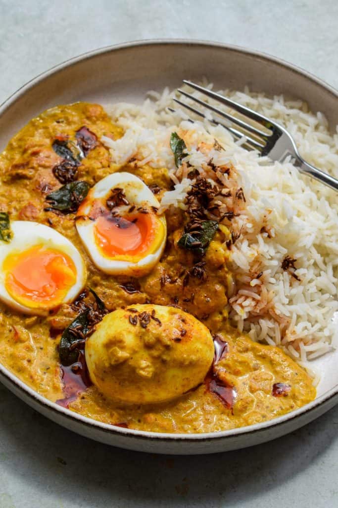 Close up of a plate of boiled egg curry and rice. One egg is left whole and the other is cut in half to show a runny middle.
