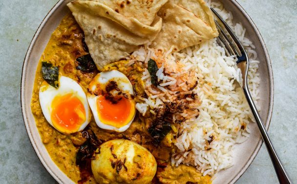 Bowl of egg curry, rice and roti close up. One of the eggs is cut in half to show the soft middle.