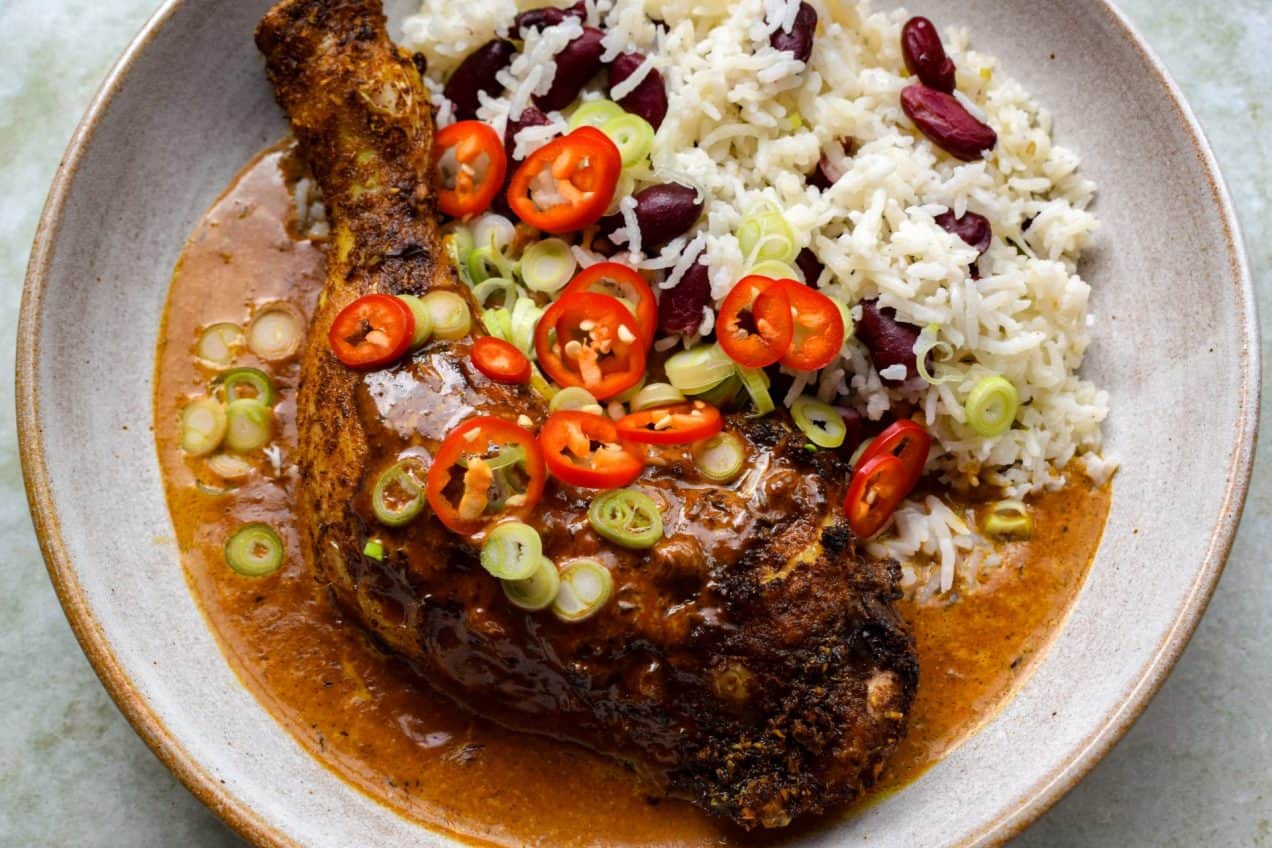 Jerk chicken leg in cream sauce with red beans and rice. Chopped spring onions and chillies are scattered on top.