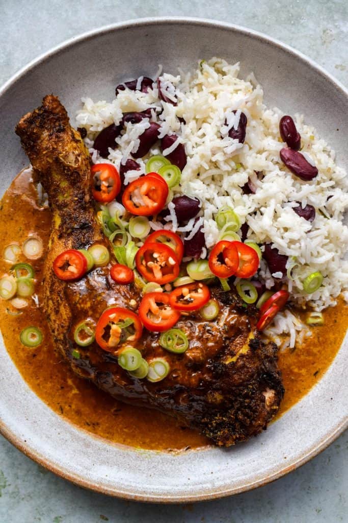 Close up of jerk chicken with a cream gravy, red beans and rice, topped with sliced chillies and spring onions.