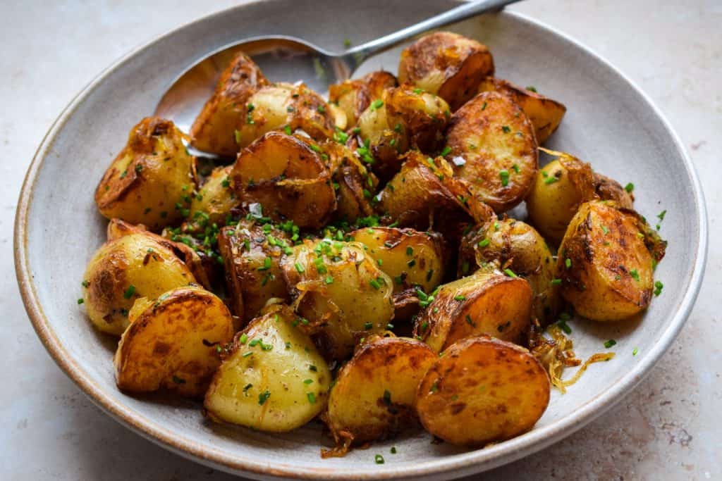 Side-on view of a bowl of fried new potatoes topped with caramalised onions and chopped chives. 