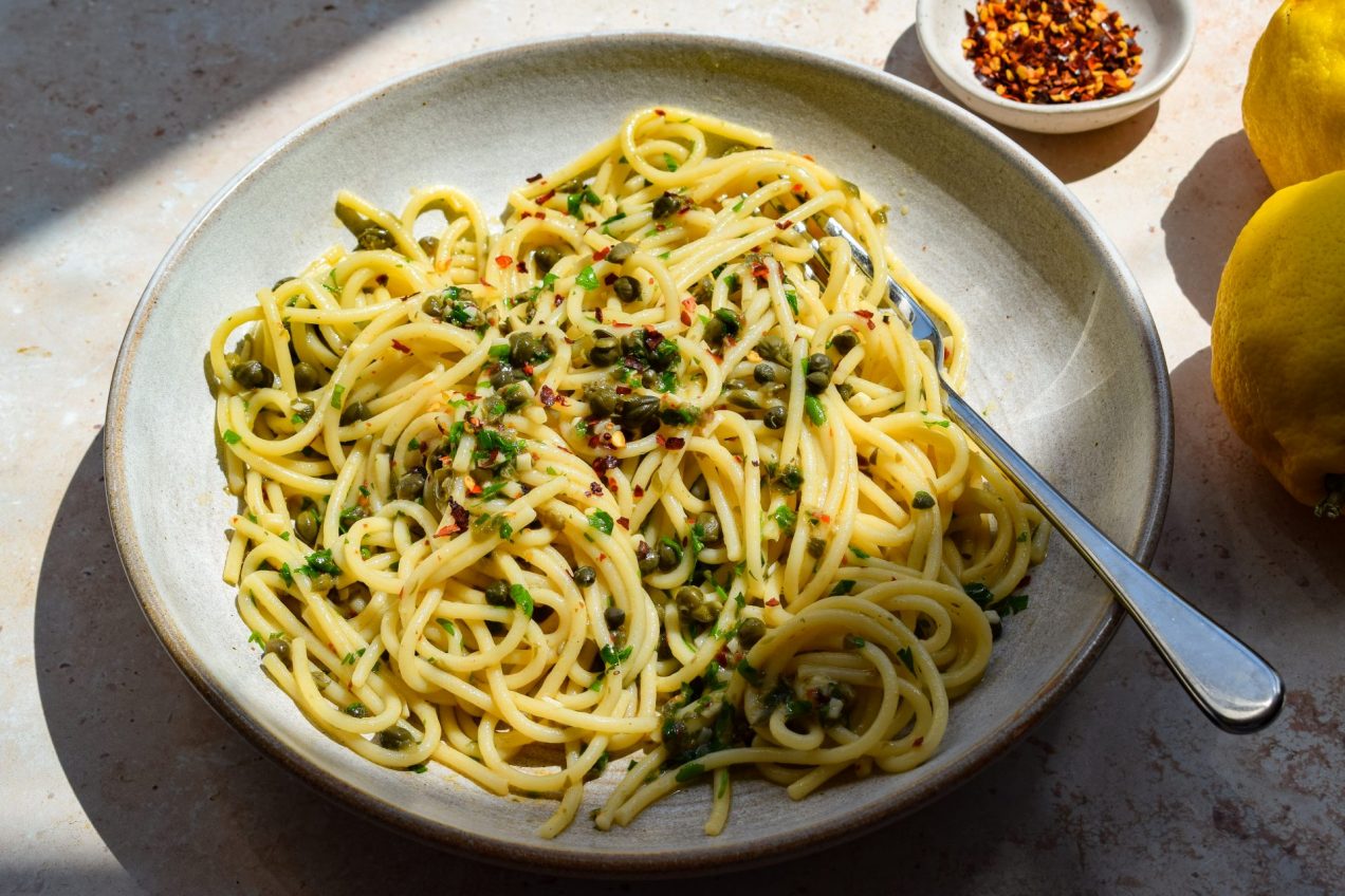Side-on view of a stone bowl of spaghetti with capers and chilli, with a bowl of chilli flakes and whole lemons off to one side.