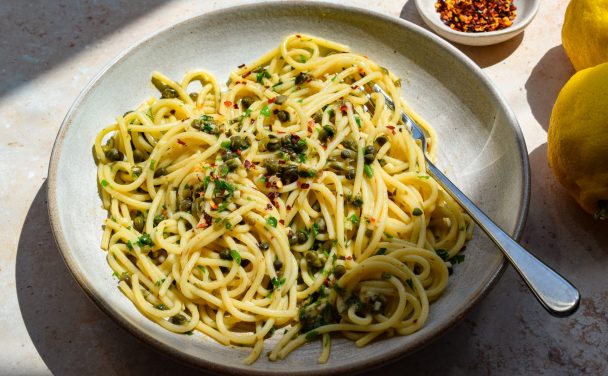 Side-on view of a stone bowl of spaghetti with capers and chilli, with a bowl of chilli flakes and whole lemons off to one side.