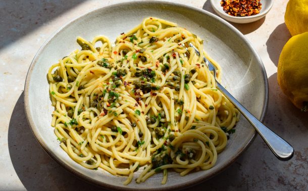 Side-on view of a stone bowl of spaghetti with capers and chilli, with a bowl of chilli flakes and whole lemons off to one side.