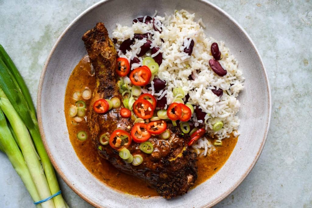 A bowl of Jerk chicken, rice, beans, chillies and spring onion slices with a cream sauce. A bunch of spring onions is sitting off to one side.