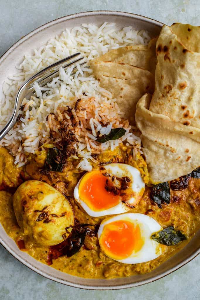 Close up of a plate of yellow curry, boiled eggs (one split in half to show the orange yolk) and rice topped with a tadka drizzle.