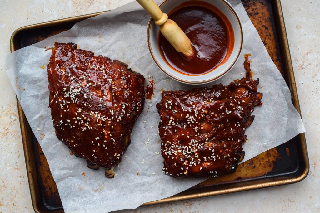 Baking sheet with two half racks of ribs and a bowl of gochujang barbecue sauce.