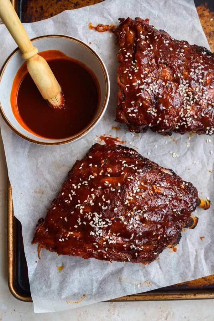 A rack of ribs divided in half with sesame seeds and gochujang barbecue sauce. The bowl of sauce with a brush is off to one side.
