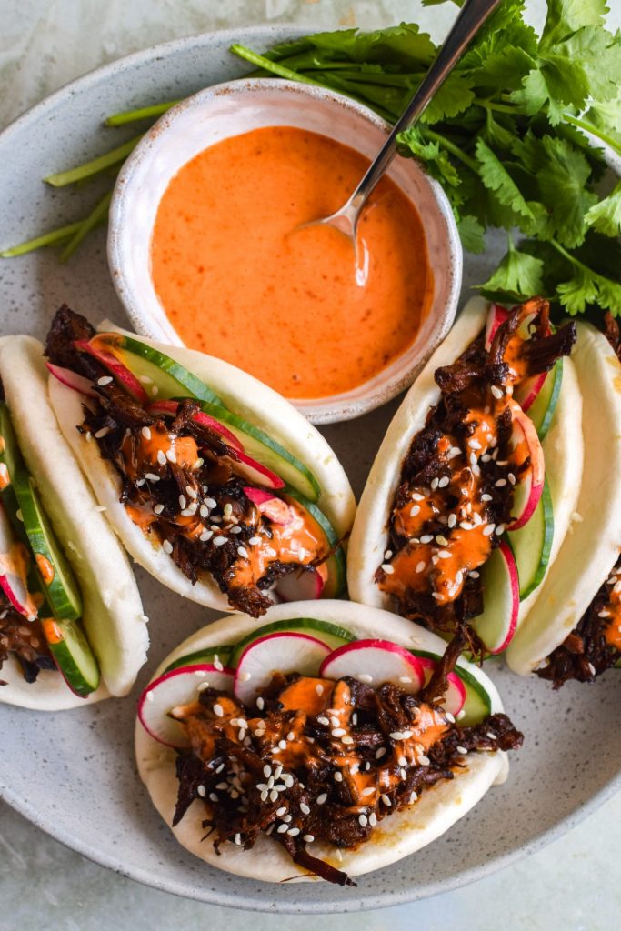 Four bao buns on a plate with a bowl of spicy creamy sauce.