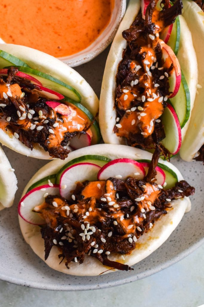 Close up of 3 hoisin pulled pork bao buns with sliced cucumber, radishes, gochujang drizzle and sesame seeds.