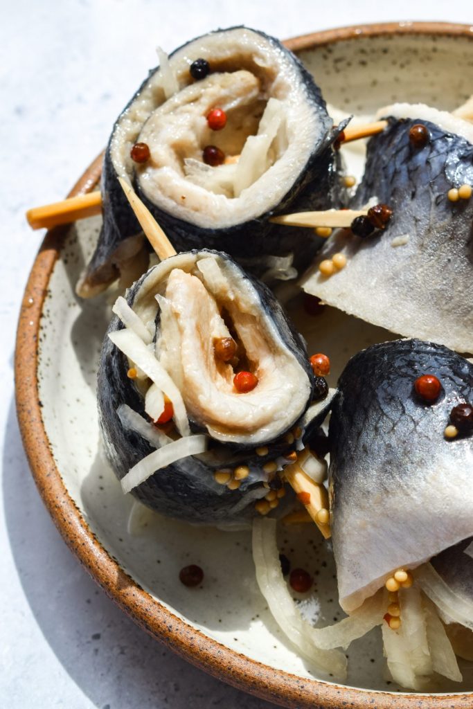 Close up of rollmops with onion, red peppercorns and yellow mustard seeds on a small plate.