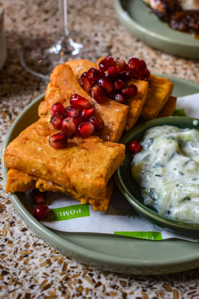 Green plate of square chickpea fritters topped with pomegranate seeds. There is also a small dish of tzatziki on the plate.
