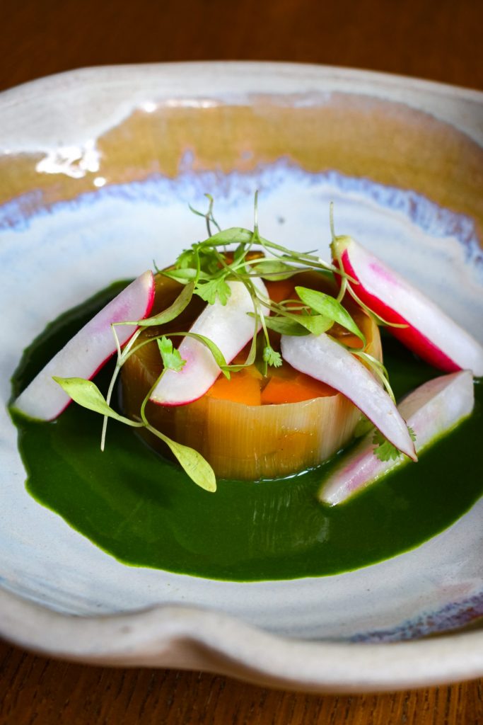 Round of vegetable terrine sitting in a puddle of green sauce topped with sliced radishes and micro herbs.