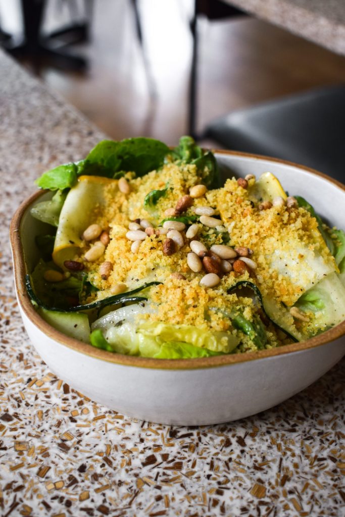 Small bowl of courgette ribbon and little gem salad topped with pine nuts and a yellow crumb.