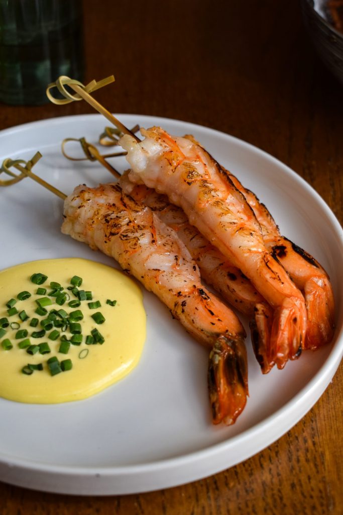 Pile of prawn tails on skewers on a white plate with a puddle of chive-topped saffron aioli. 