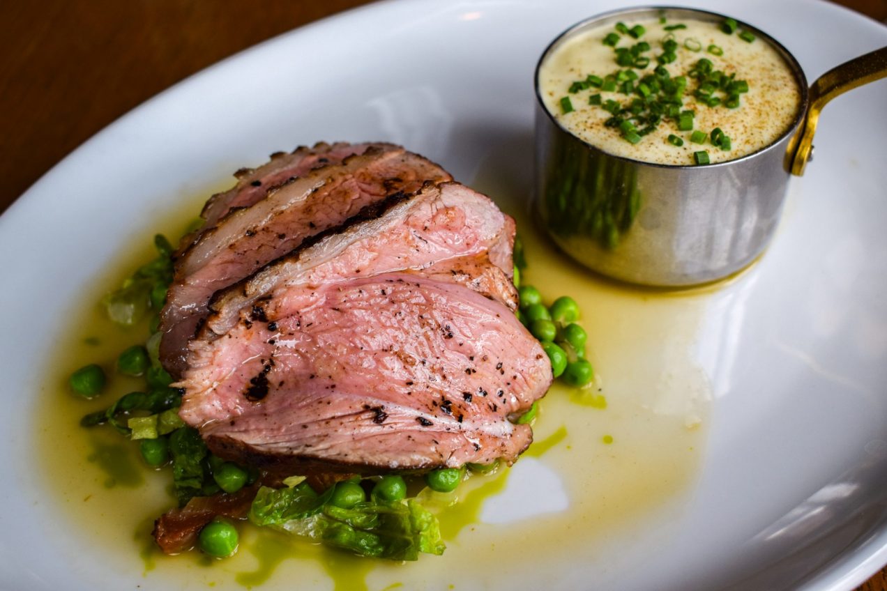 Slices of lamb rump on a pile of peas a la Francaise with a small pan of lamb pie topped with chives in the background.