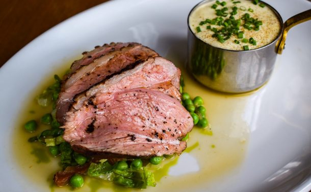 Slices of lamb rump on a pile of peas a la Francaise with a small pan of lamb pie topped with chives in the background.
