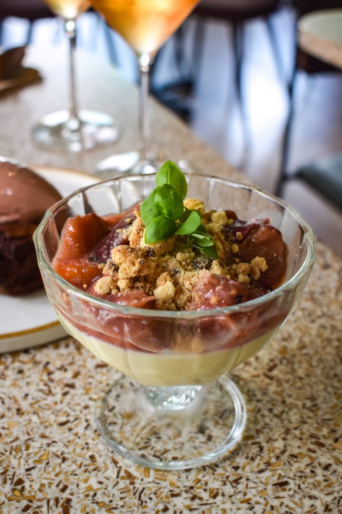 Lemon posset topped with a peach and raspberry compote, biscuit crumbs and basil leaves in a glass serving dish.