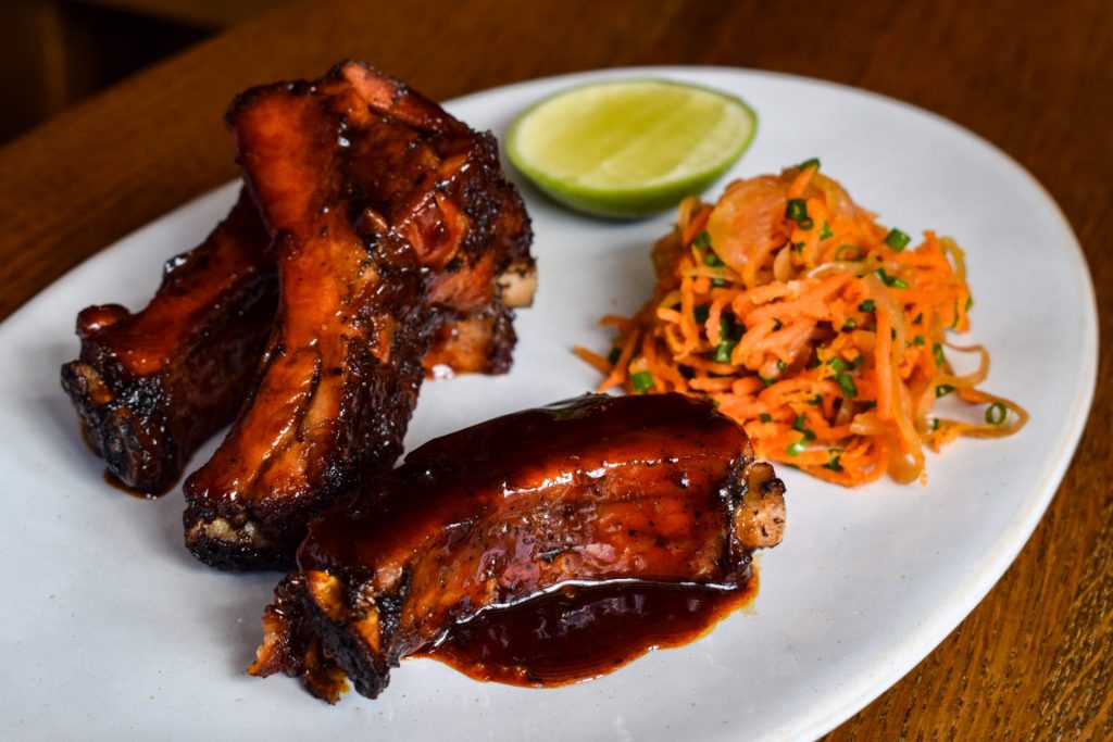 Three glazed pork ribs on a white plate next to a tangle of carrot slaw and a lime wedge.