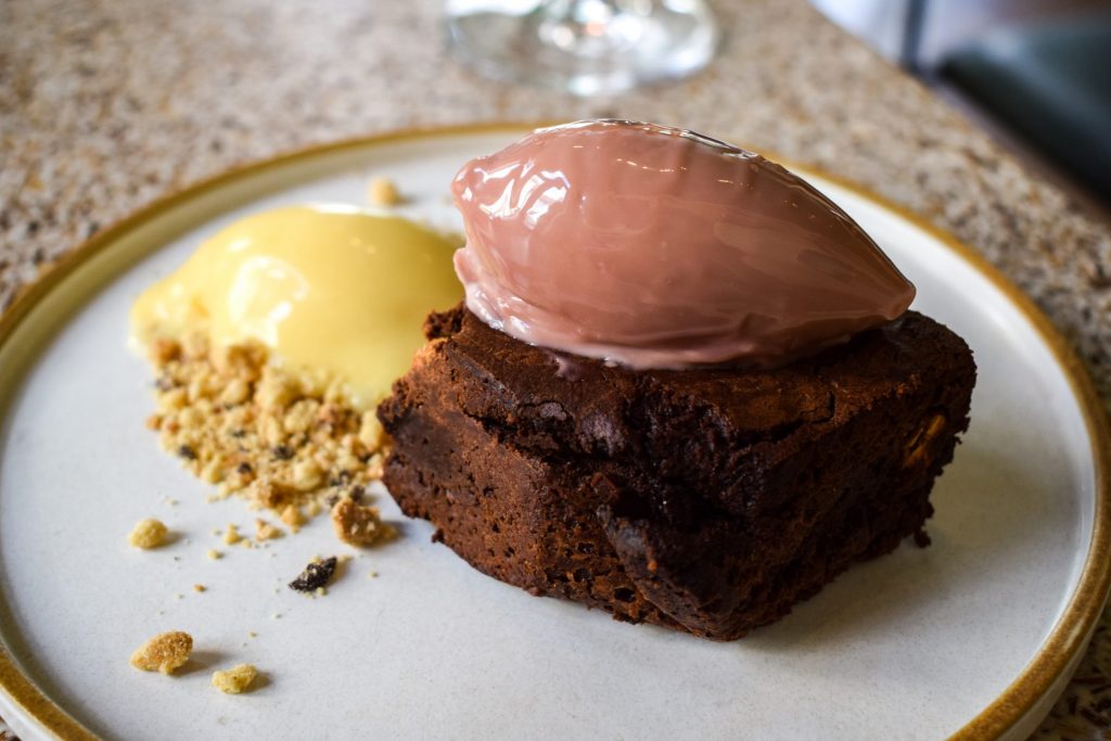 Wedge of parkin on a plate topped with a quenelle of ruby chocolate with another of custard and biscuit crumbs next to it on the plate.