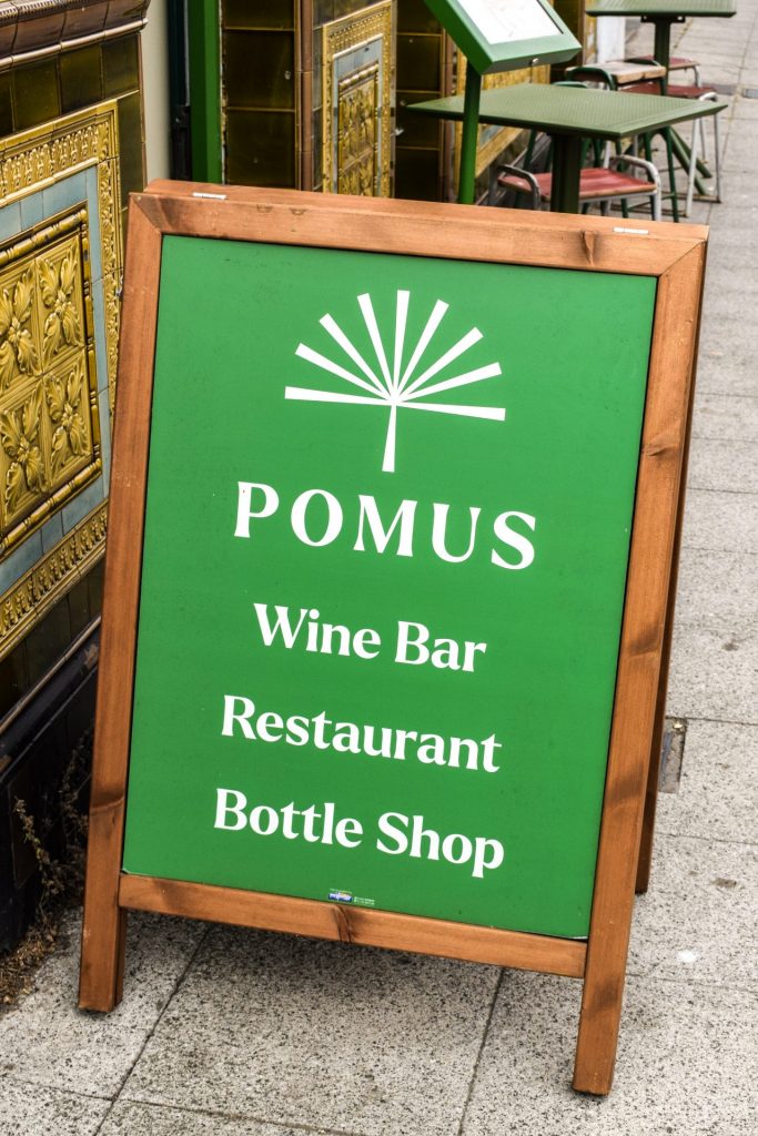 Green sandwich board advertising that Pomus, Folkestone is a wine bar, restaurant and bottle shop in the street outside.
