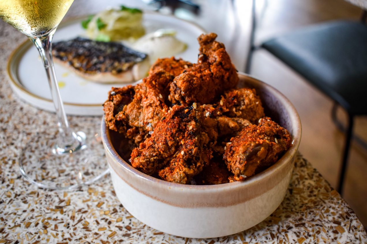Bowl of chilli dusted fried popcorn chicken, with a plate of mackerel and the stem of a glass of white wine in the background.