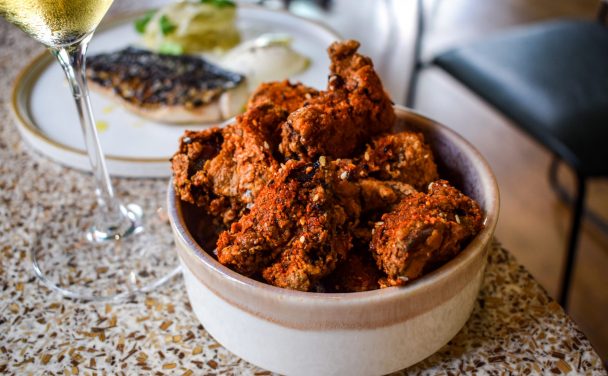 Bowl of chilli dusted fried popcorn chicken, with a plate of mackerel and the stem of a glass of white wine in the background.