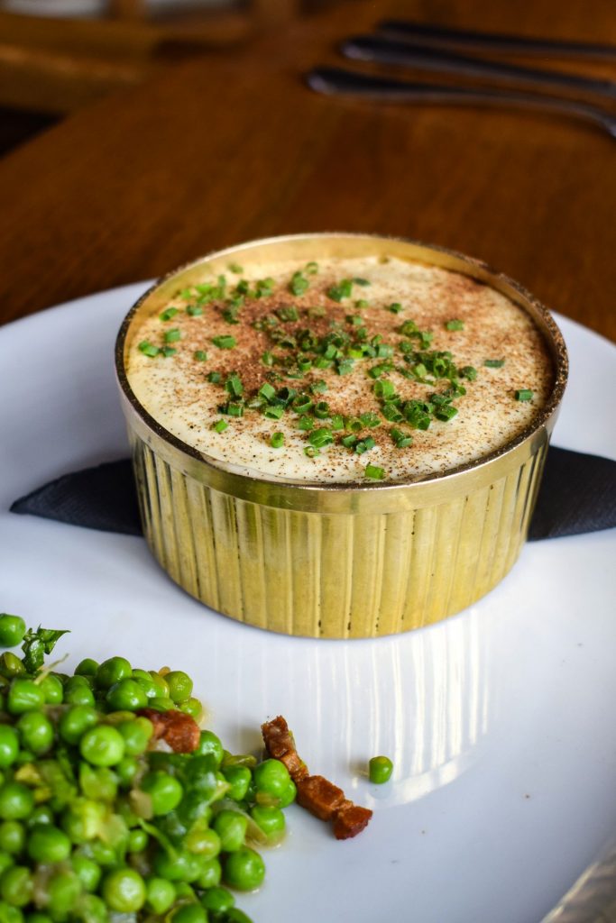 Lamb pie topped with chives in a gold dish on a white plate. There is a pile of peas and bacon in the foreground.
