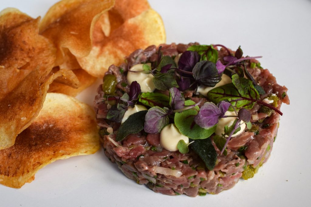 Steak tartare topped with mayo and micro herbs next to a pile of homemade chips.