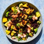 Bowl of black rice salad with prawns, avocado, mango, and Tajin sprinkled on top on a blue background.