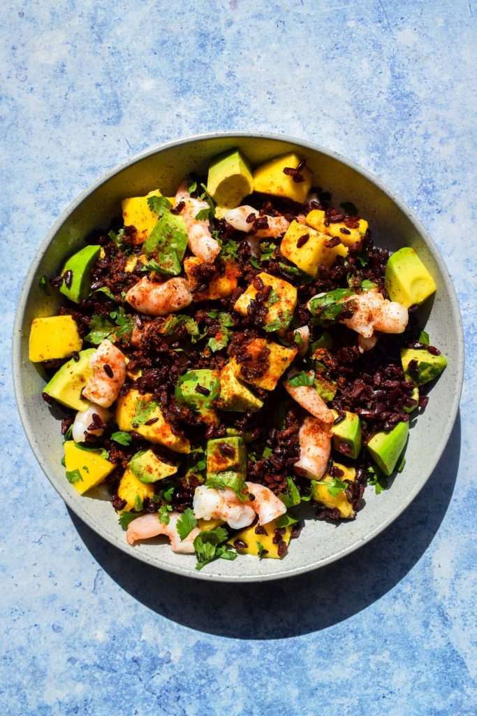 Bowl of black rice salad with prawns, avocado, mango, and Tajin sprinkled on top on a blue background.