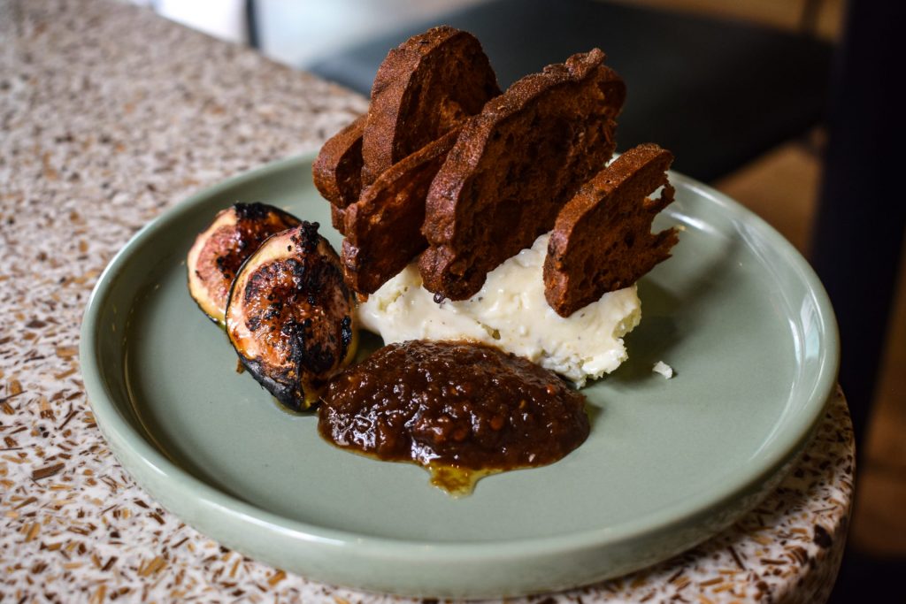A green plate of whipped goats cheese, fig jam and torched fig halves with crackers standing up in the goats cheese.