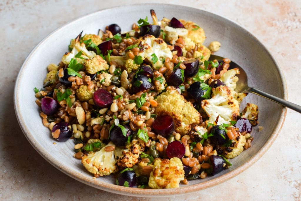 Side on view of a grain salad with parsley, roasted cauliflower, nuts, seeds and grapes.