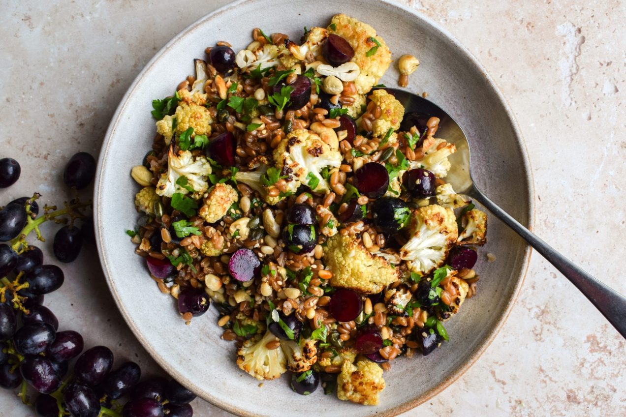 Grape and roasted cauliflower grain salad in a bowl with a bunch of grapes off to the side.
