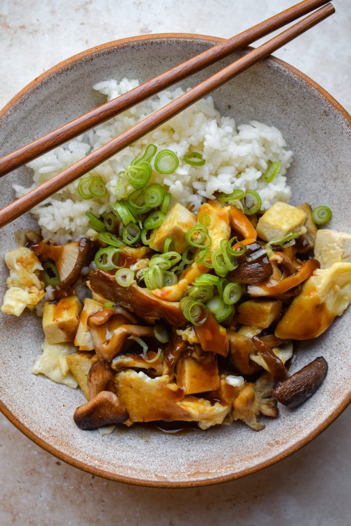 Bowl of mushroom, egg and tofu stir fry with a Japanese gravy served with rice and thinly slices spring onions. There is a set of wooden chopsticks perched on the side of the bowl.