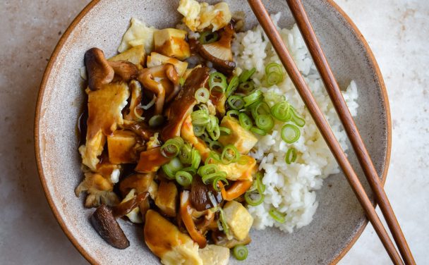 Close up of a bowl of mushroom, tofu and egg stir fry served with white rice, spring onions and a glossy ankake sauce.