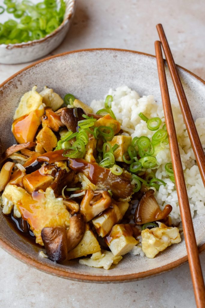Bowl of Japanese mushroom, tofu and egg stir fry with white rice, sliced spring onion and a thick glossy sauce. There is a bowl of sliced spring onions also in the background.