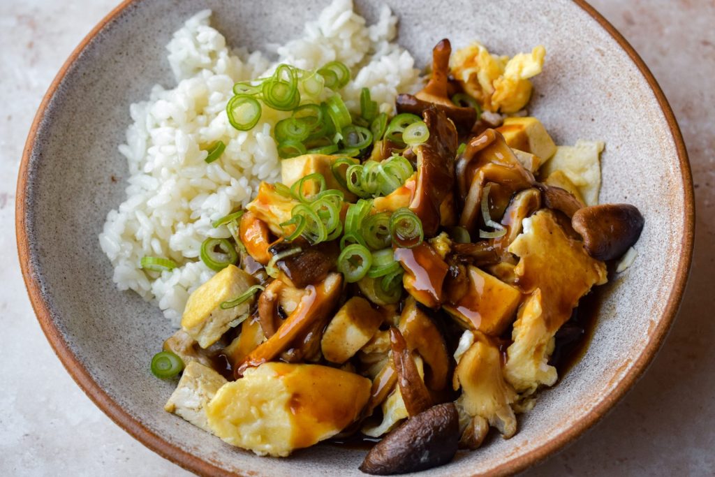 Close up of oyster mushrooms and shiitake stir fried with egg and tofu, served with rice, sliced spring onions and a thick, glossy brown sauce.