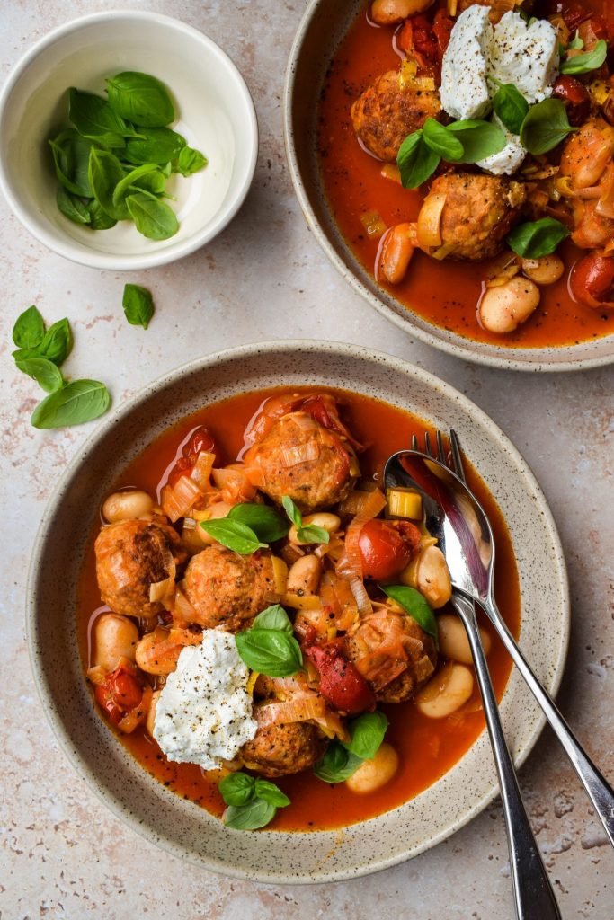 Two bowls of turkey meatballs with tomatoes, butter beans and ricotta. They're scattered with basil leaves with an extra bowl of them off to one side.